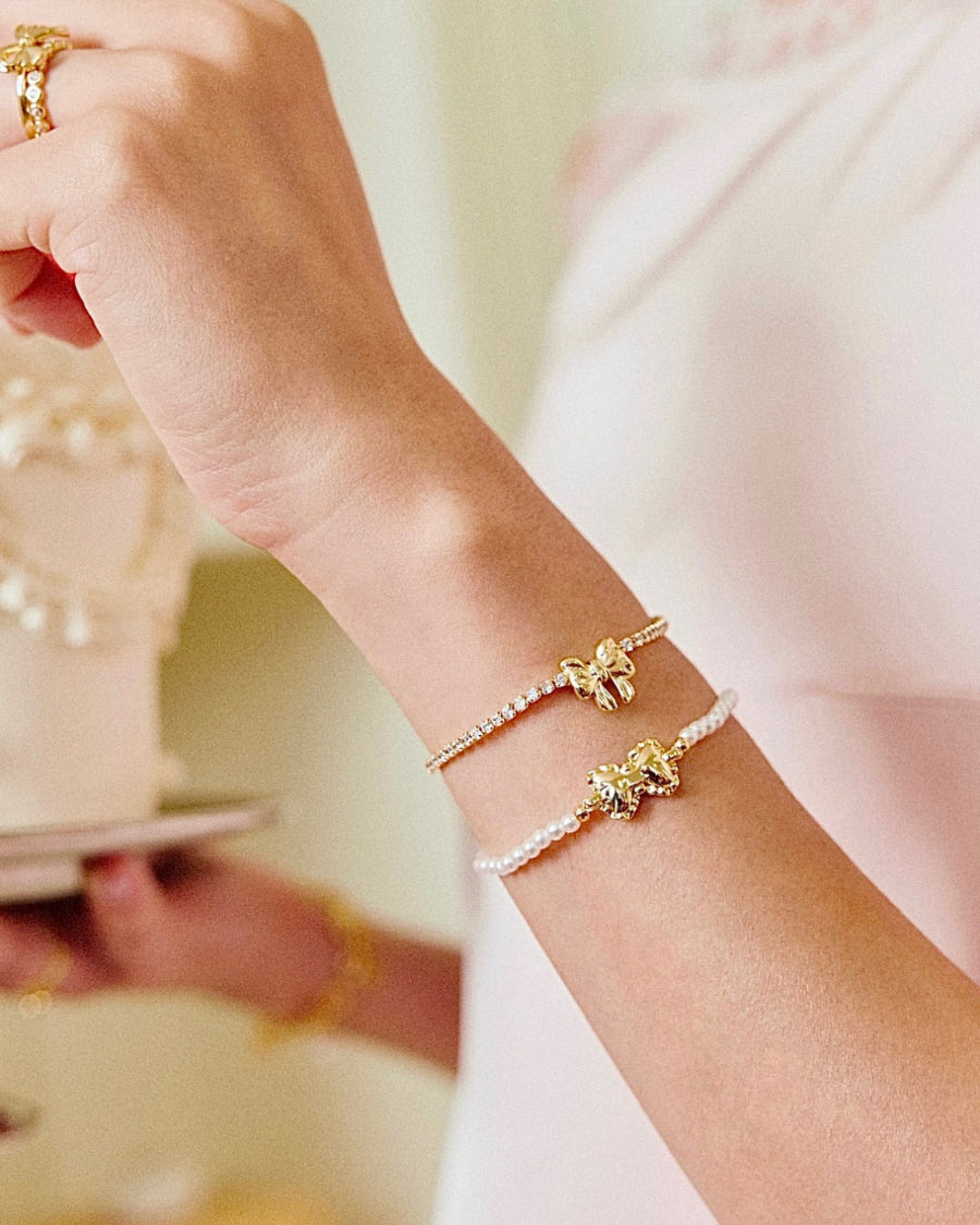 Close-up of a wrist wearing two gold bracelets with floral designs, against a blurred background.